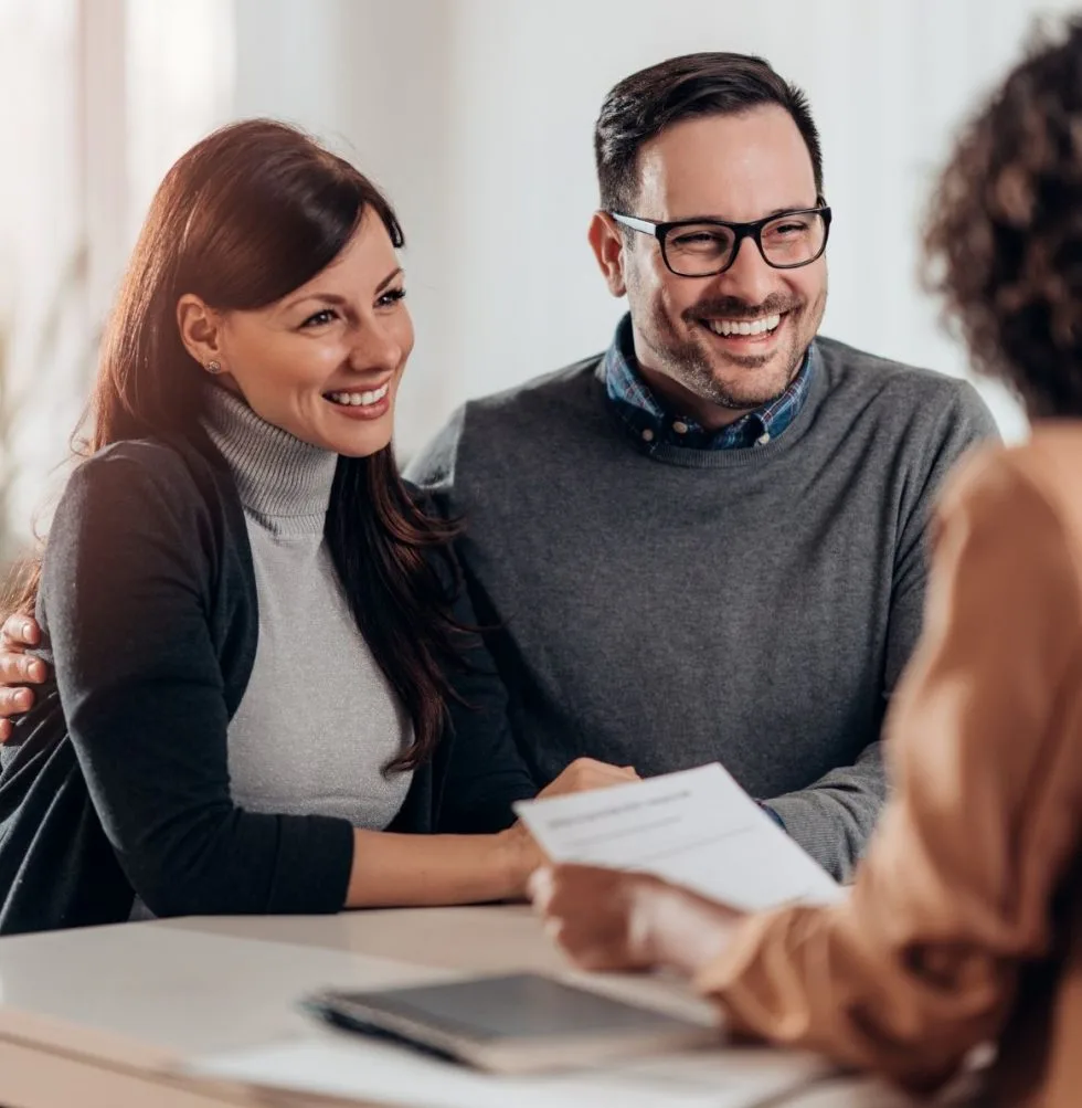 Un couple souriant échange avec un agent immobilier, assis à une table de bureau